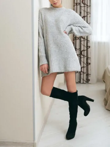 Girl wearing beige sweater dress with boots that are black and knee high, leaning against bedroom wall.