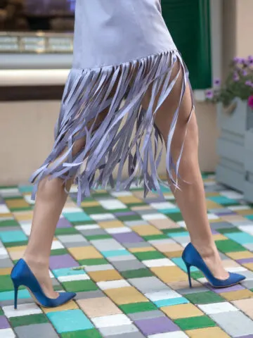 Close up of women's legs wearing blue heeled shoes with skirt that is fringed and purple walking on colorful cobblestones.