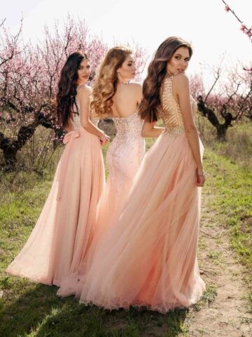 3 women wearing peach dresses and gowns walking in cherry blossom field.