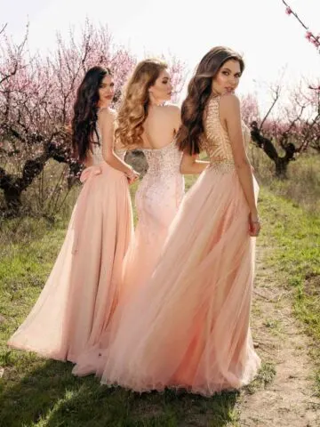 3 women wearing peach dresses and gowns walking in cherry blossom field.