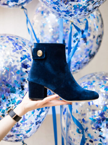 Woman's hand holding blue velvet ankle boot in front of balloons.
