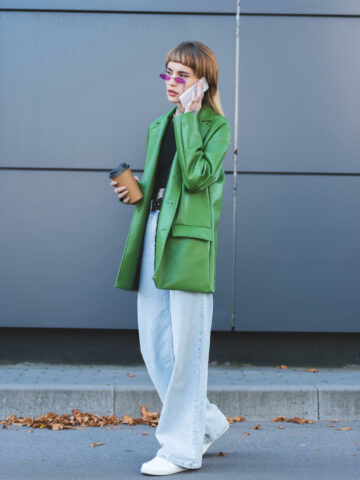 Woman wearing white sneaker shoes with baggy jeans green leather jacket and purple sunglasses talking on her phone.