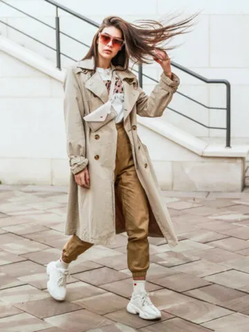 Woman walking wearing tan joggers with sneaker shoes, beige trench and red sunglasses and flipping her hair.