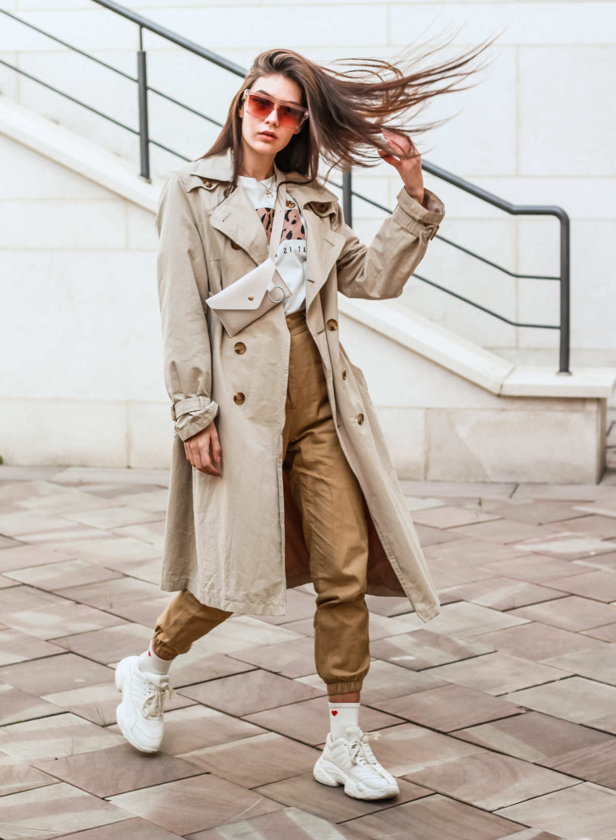 Woman walking wearing tan joggers with sneaker shoes, beige trench and red sunglasses and flipping her hair.
