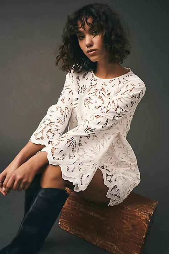 Short haired brunette woman sitting on piece of wood wearing a lace dress with cowboy boots.