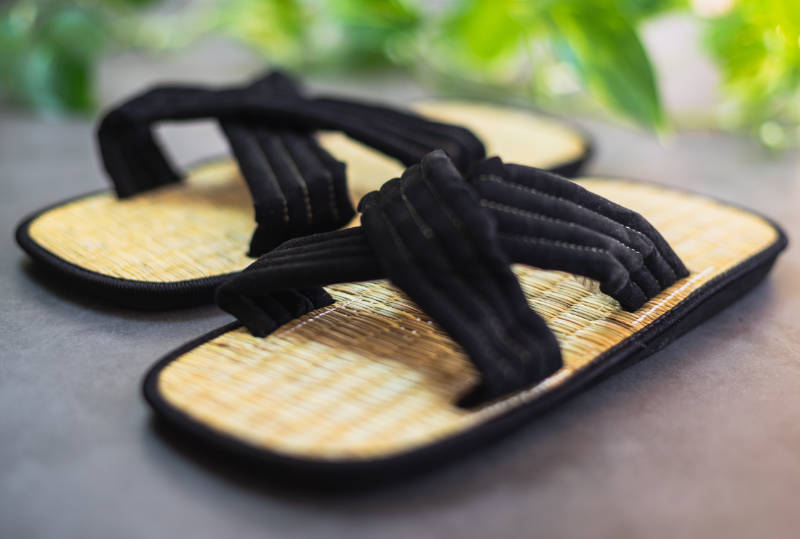 Pair of tatami sandals with black criss cross straps on a grey table with leaves in the background.