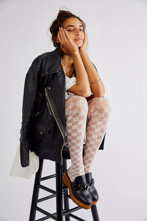 Woman sitting on bench wearing clogs with tights and a black moto jacket.