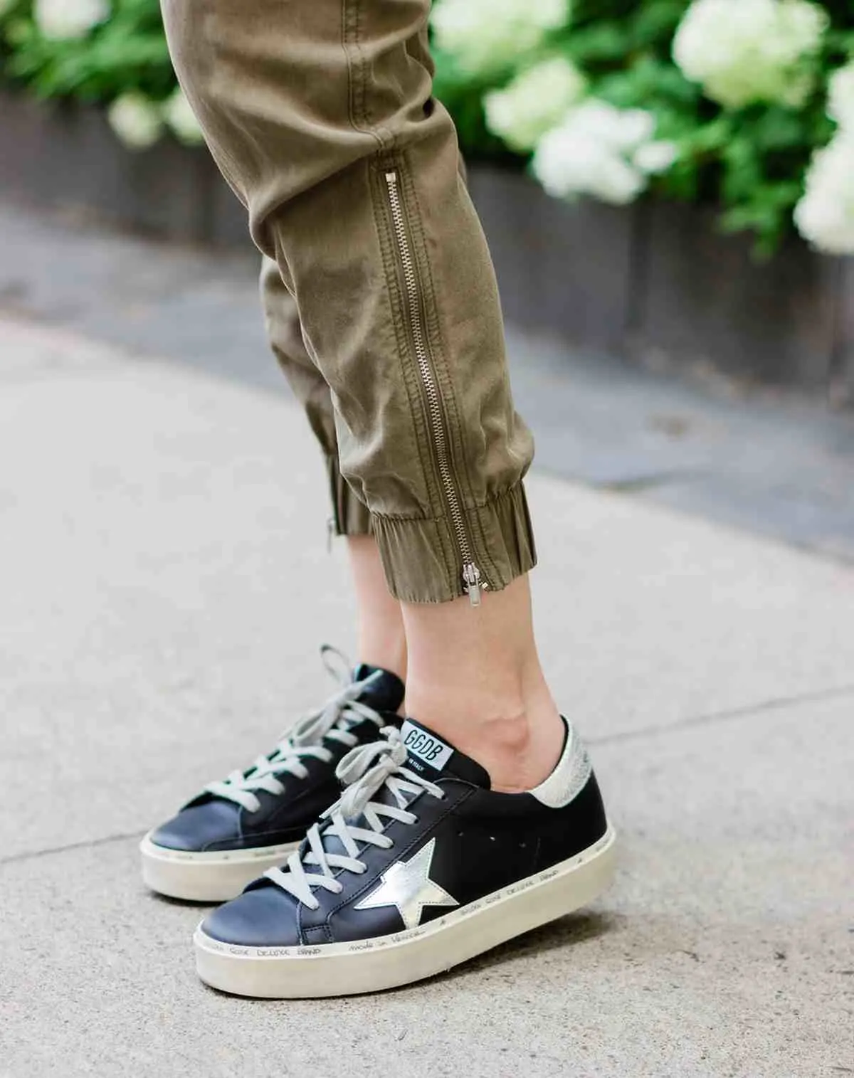 Close up of woman's feet wearing black golden goose star sneakers and khaki pants on a woven chair on concrete sidewalk with white and green flowers.