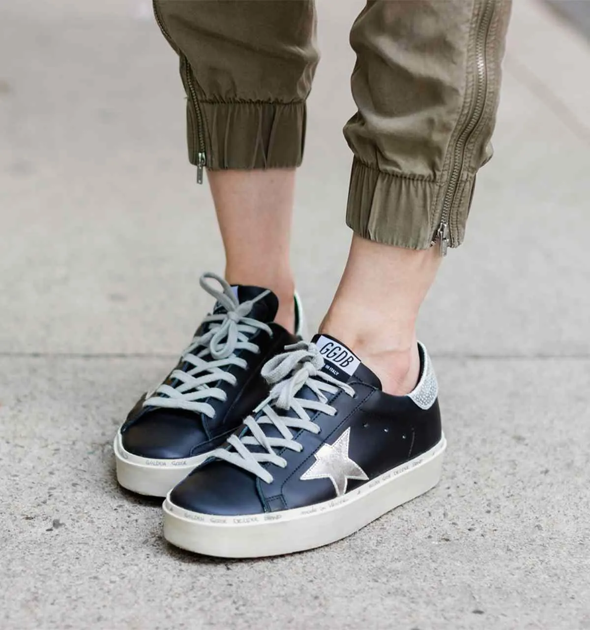 Close up of woman's feet wearing black golden goose star sneakers and khaki pants on a woven chair standing on concrete sidewalk.