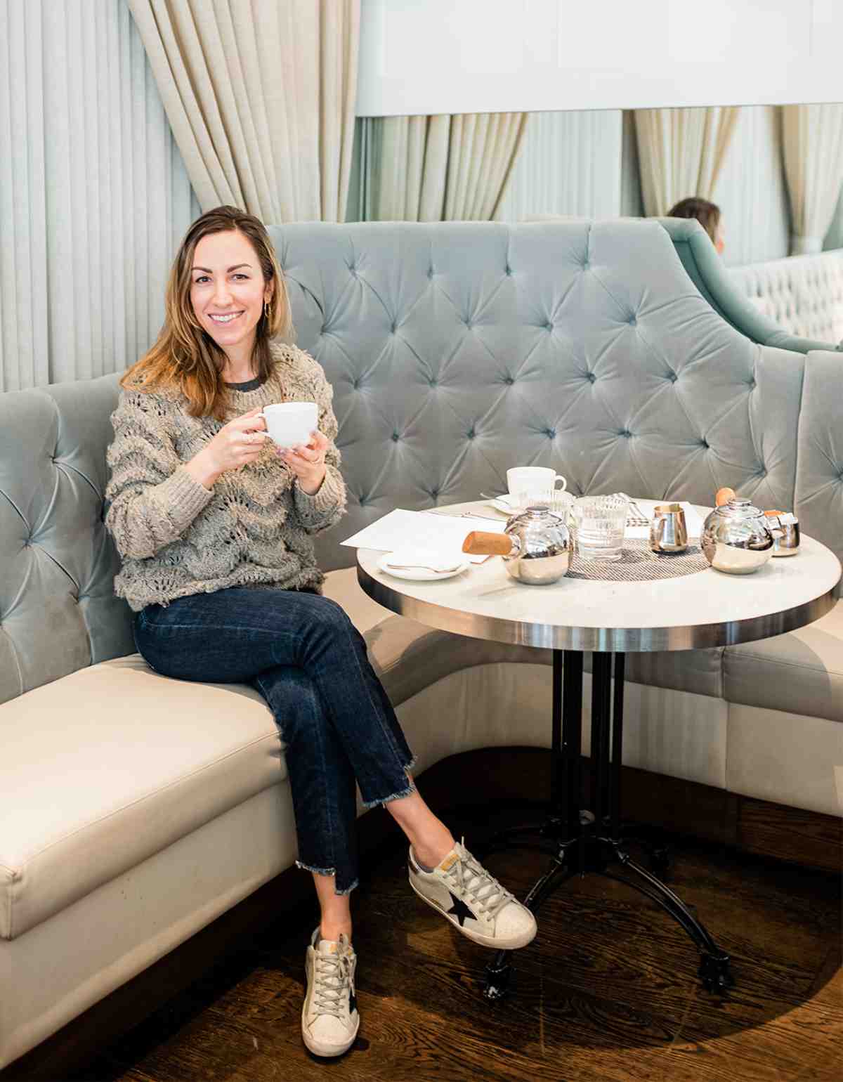Smiling blonde woman sitting in booth of cafe holding a tea cup wearing jeans golden goose star sneakers and a sweater.