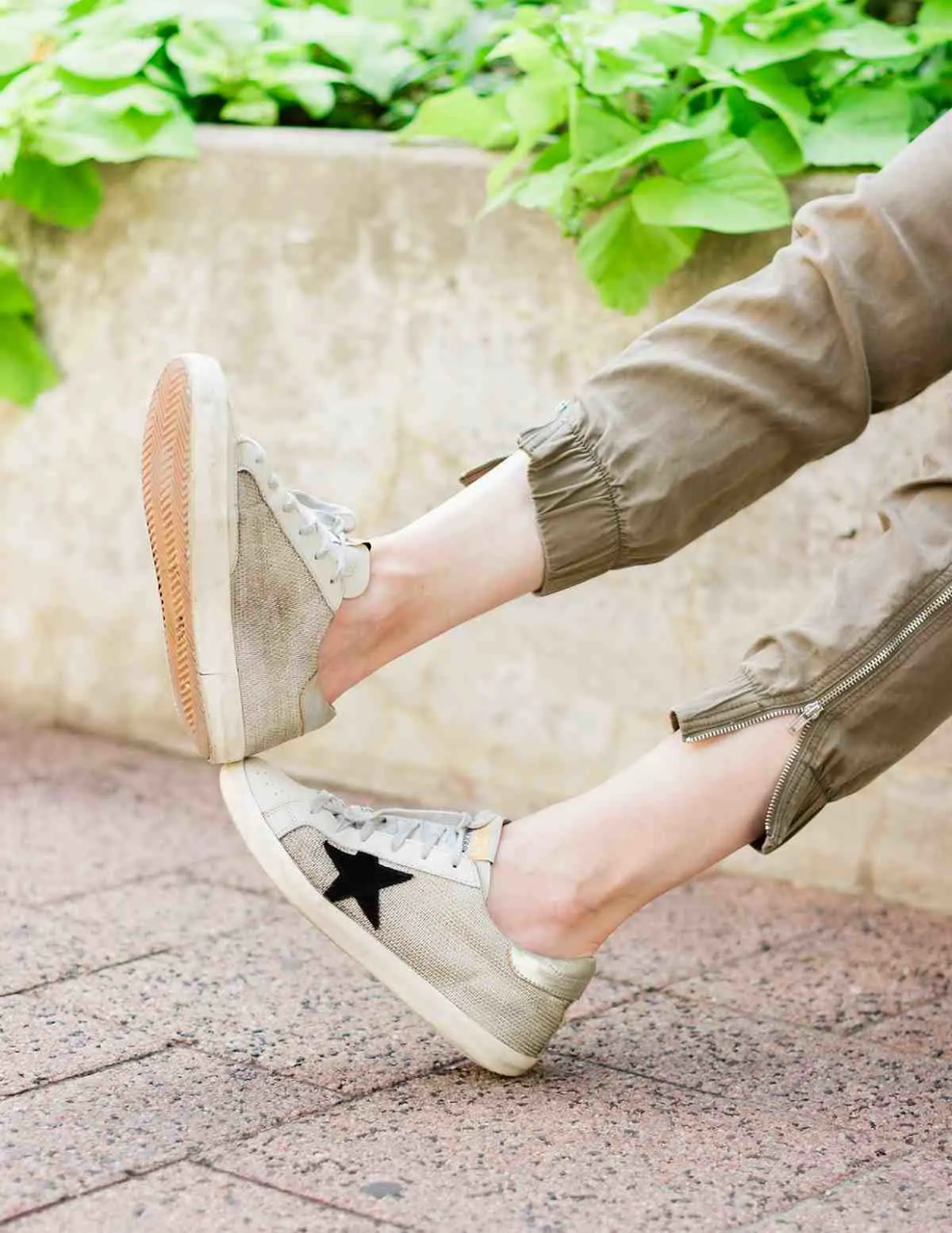 Close up of woman's feet wearing off-white golden goose star sneakers and khaki pants on a woven chair with purple concrete and leaves in the background.