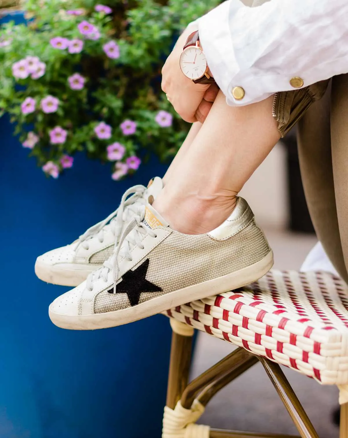 Close up of woman's feet wearing off-white golden goose star sneakers on a woven chair with purple flowers in the background.