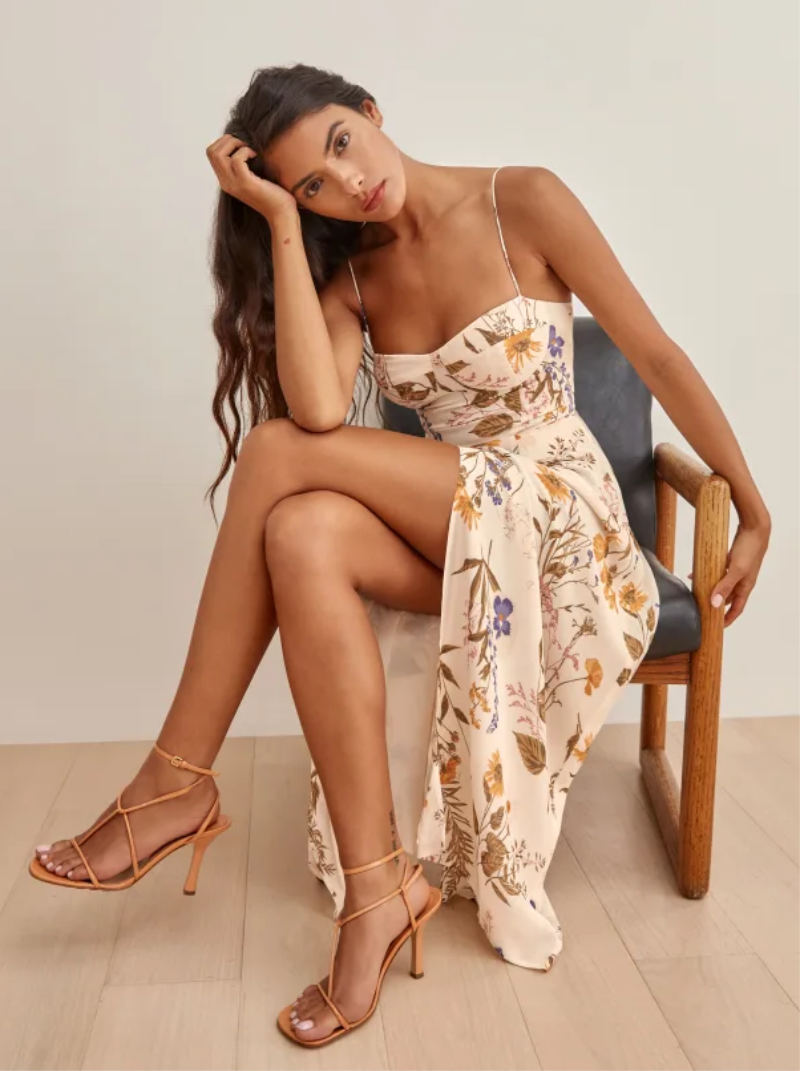 Woman sitting on chair wearing orange strappy shoes with a sundress in beige tone and floral motif.