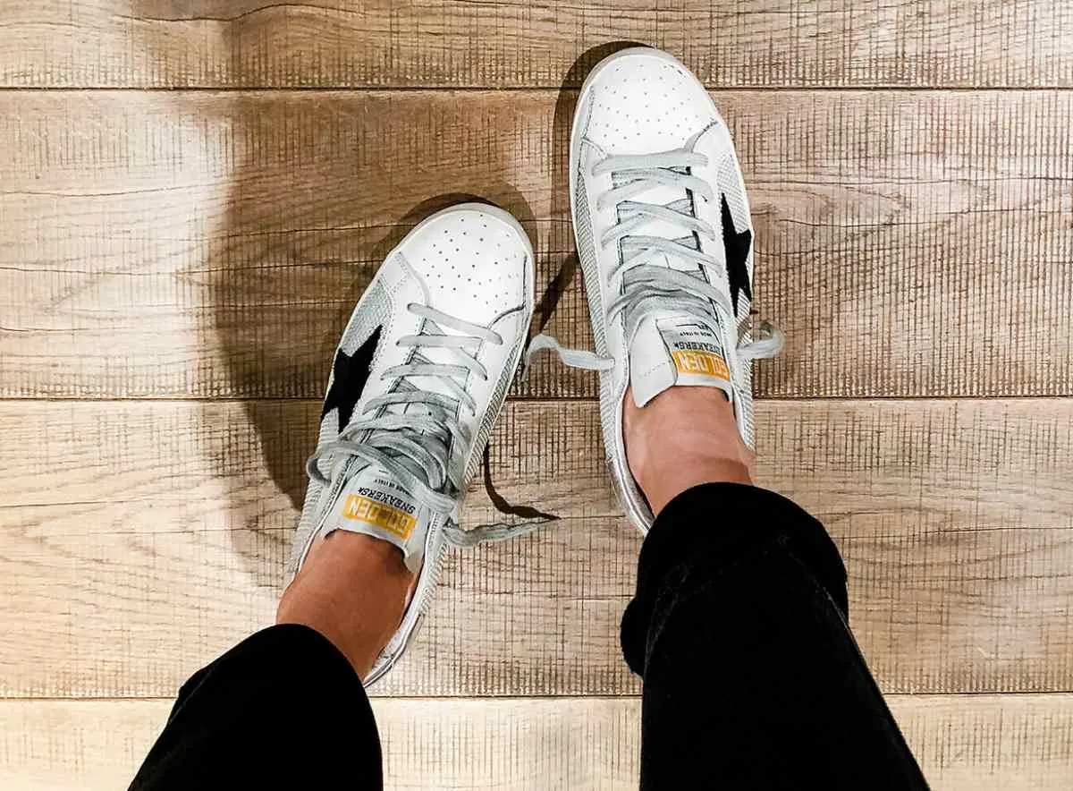 Top view of woman's feet wearing white golden goose superstar sneakers on wooden floor.
