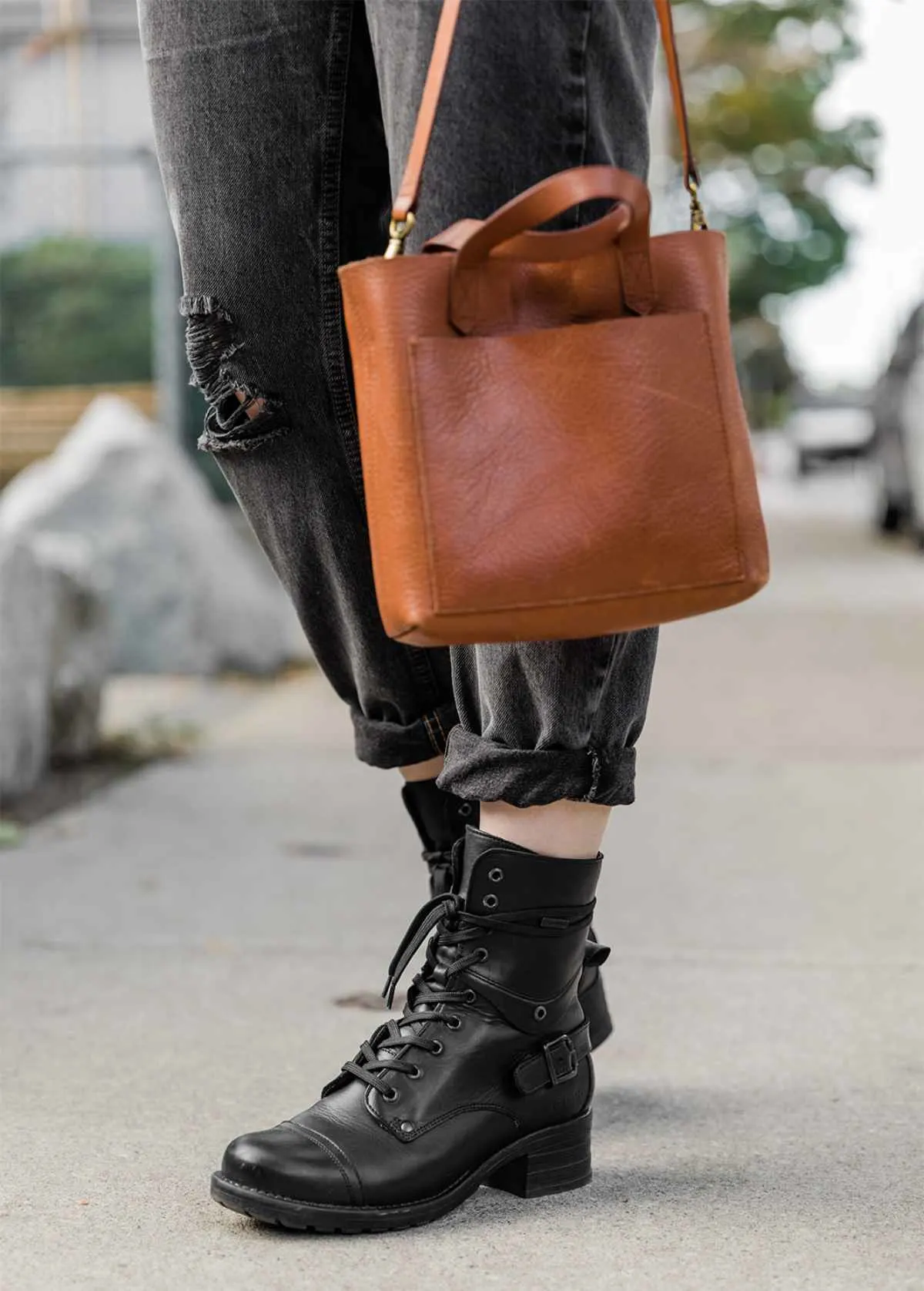 Cropped view of woman's legs wearing Taos crave combat boots in black waterproof with cuffed grey ripped jeans and a brown leather purse on a sidewalk.