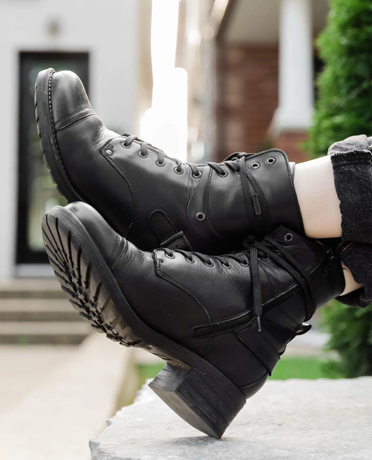 Cropped view of woman's crossed feet wearing Taos crave combat boots in black waterproof with cuffed grey jeans on a sidewalk.