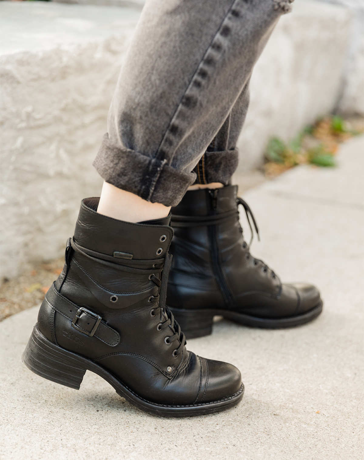 Cropped view of woman's feet wearing Taos crave combat boots in black waterproof with cuffed grey jeans on a sidewalk.