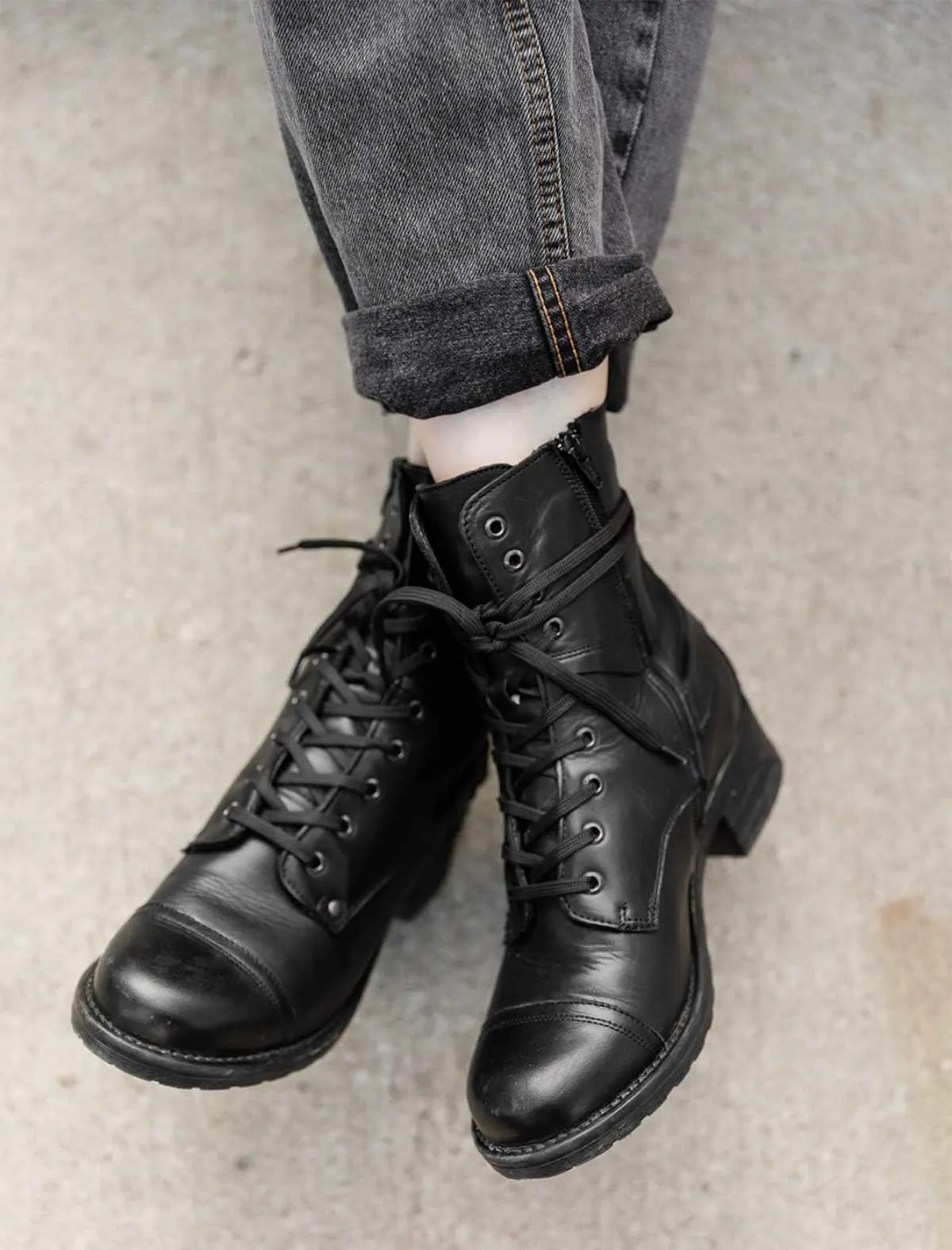 Cropped view of woman's crossed feet wearing Taos crave combat boots in black waterproof with cuffed grey jeans on a sidewalk.