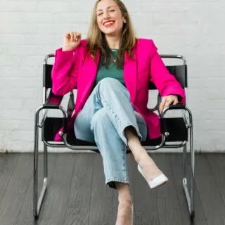 Photo of ShoeTease shoe blogger sitting in chair smiling, wearing a hot pink blazer green top faded jeans and white pumps.