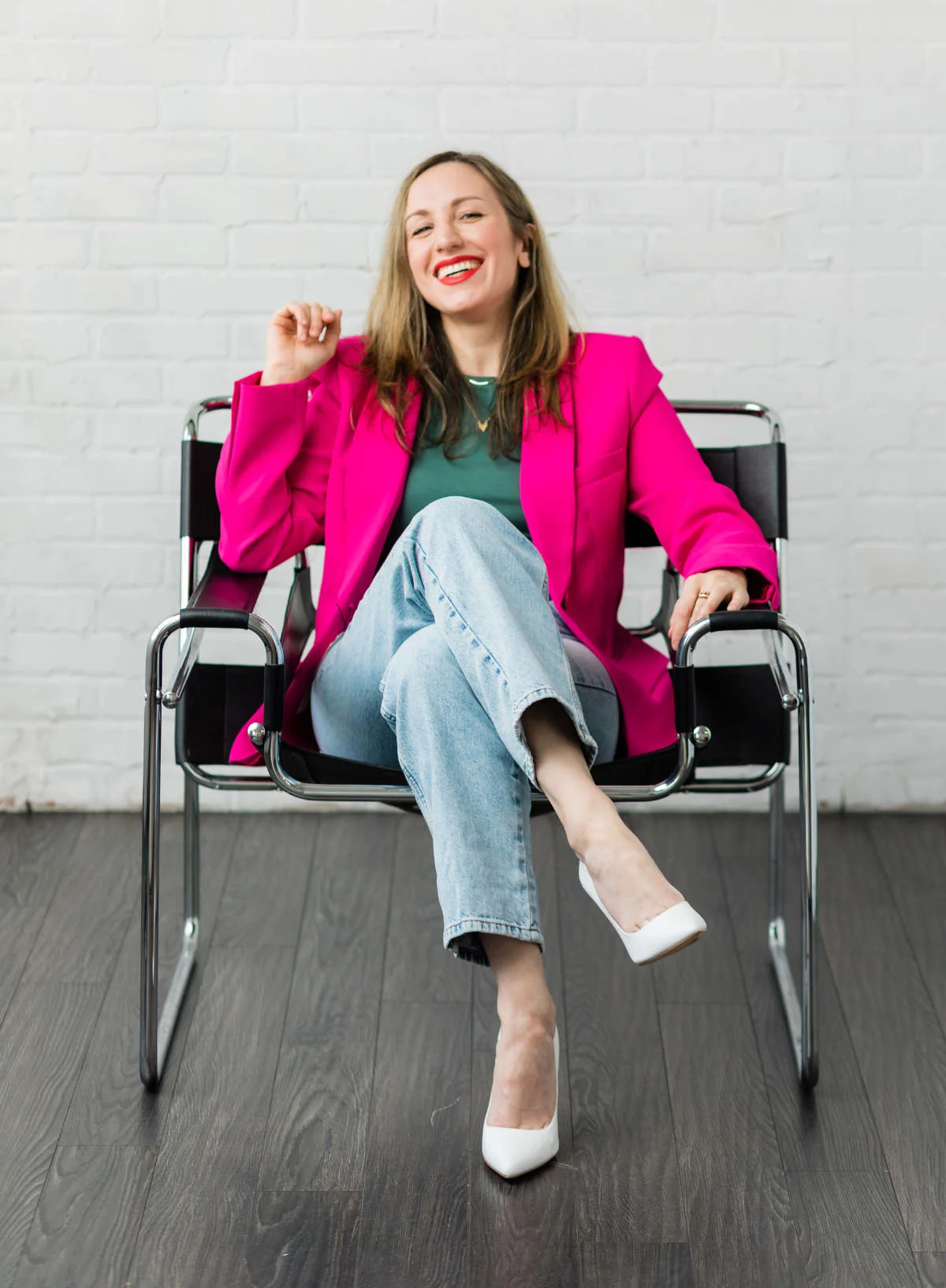 Photo of ShoeTease shoe blogger sitting in chair smiling, wearing a hot pink blazer green top faded jeans and white pumps.