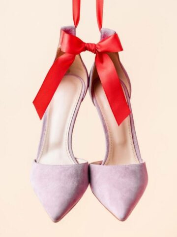 Pair of lavender suede heels hanging from a large red ribbon.