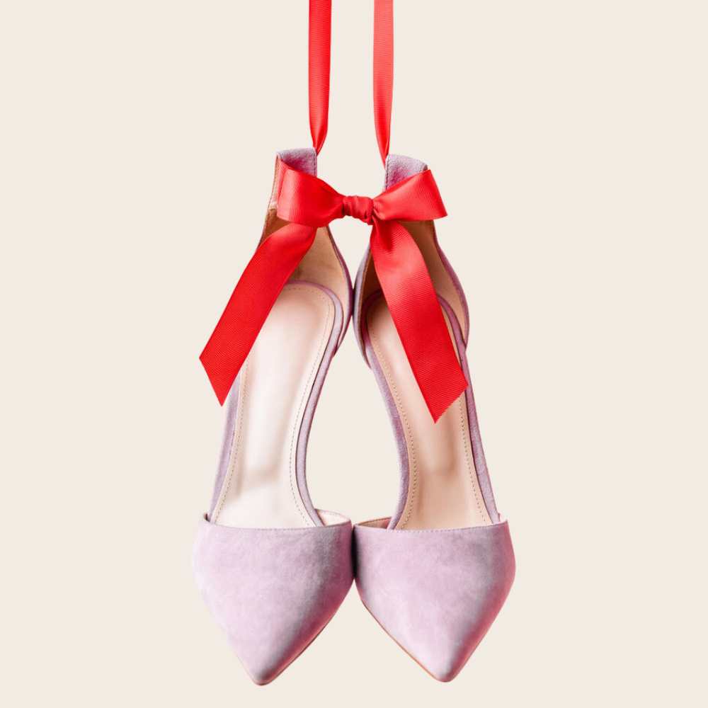 Pair of lavender suede heels hanging from a large red ribbon.