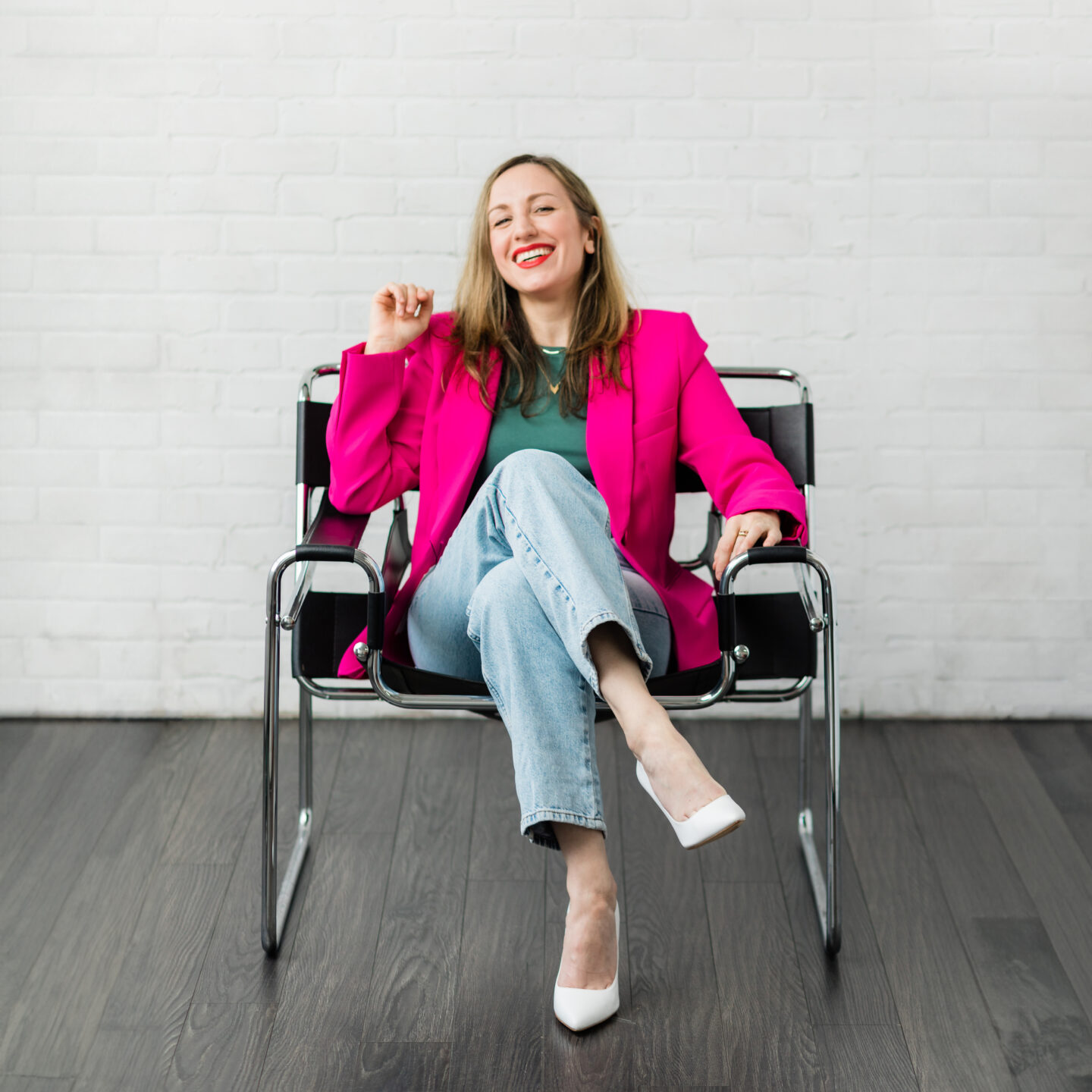 Woman sitting in a chair wearing a hot pink blazer green t shirt jeans and white heels.