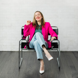 Woman sitting in a chair wearing a hot pink blazer green t shirt jeans and white heels.