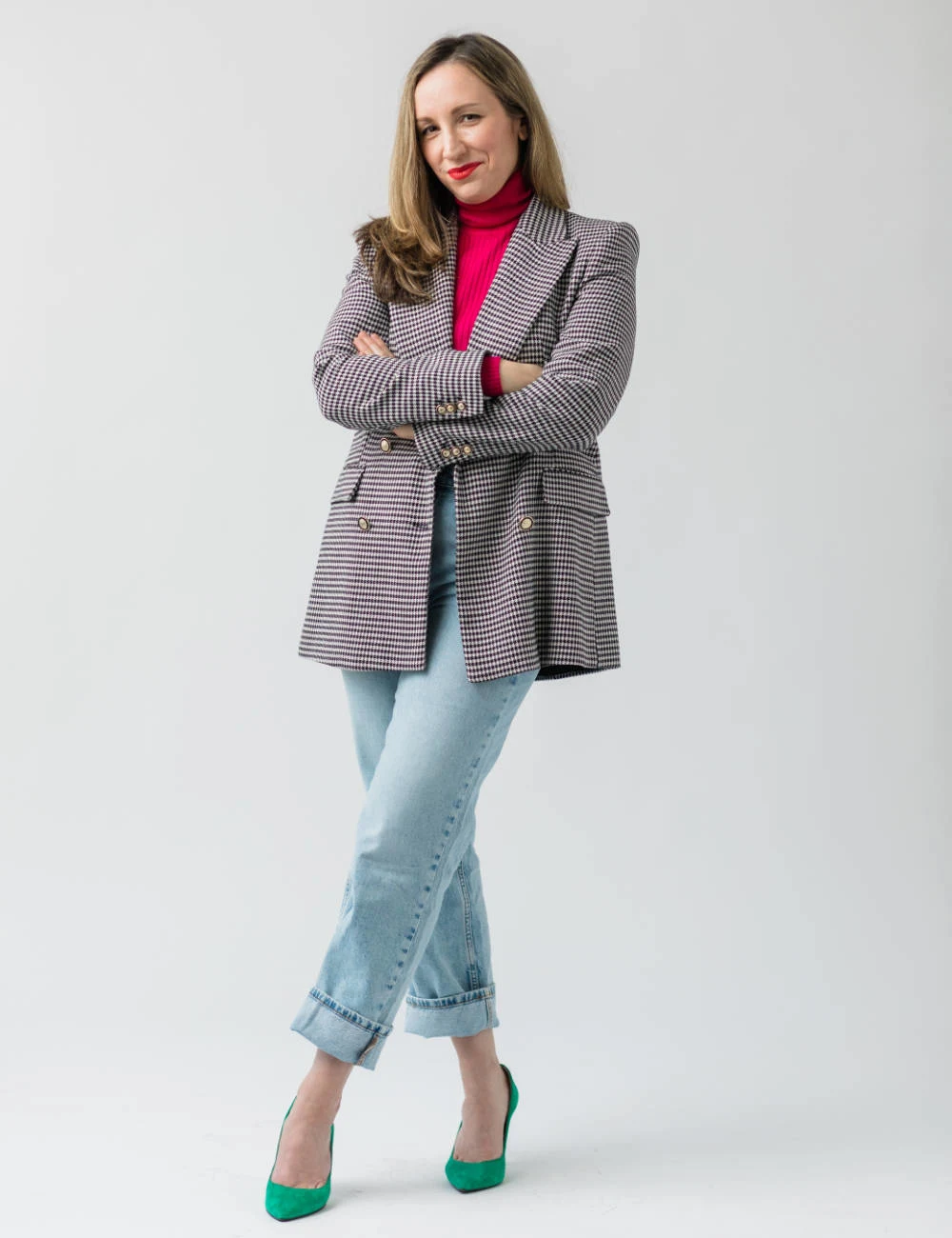 Woman of woman standing with legs and arms crossed wearing a hot pink t shirt jeans and and green heels.