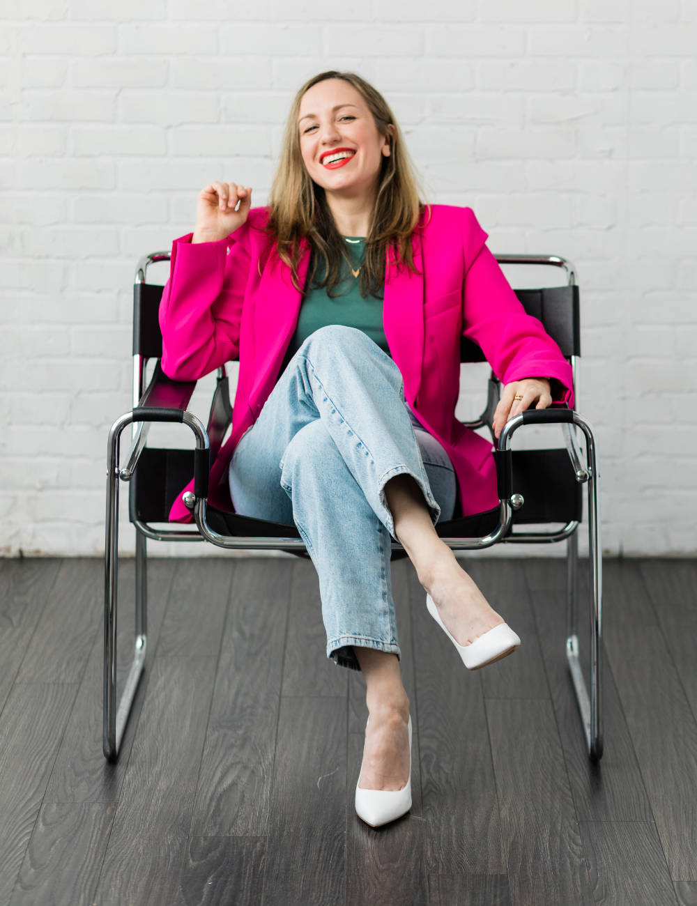 Woman sitting in a chair wearing a hot pink blazer green t shirt jeans and white heels.