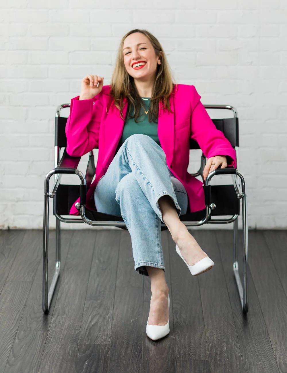 Woman sitting in a chair wearing a hot pink blazer green t shirt jeans and white heels.