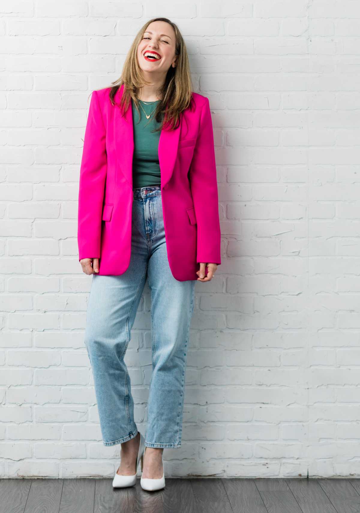 Blonde woman standing and leaning against a white brick wall wearing a hot pink blazer light blue jeans white heels and a green tshirt.