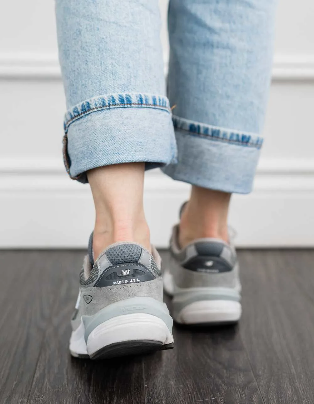 Close up of the back of a woman's feet wearing grey New Balance 990 V6 dad sneakers with rolled up faded straight leg jeans.