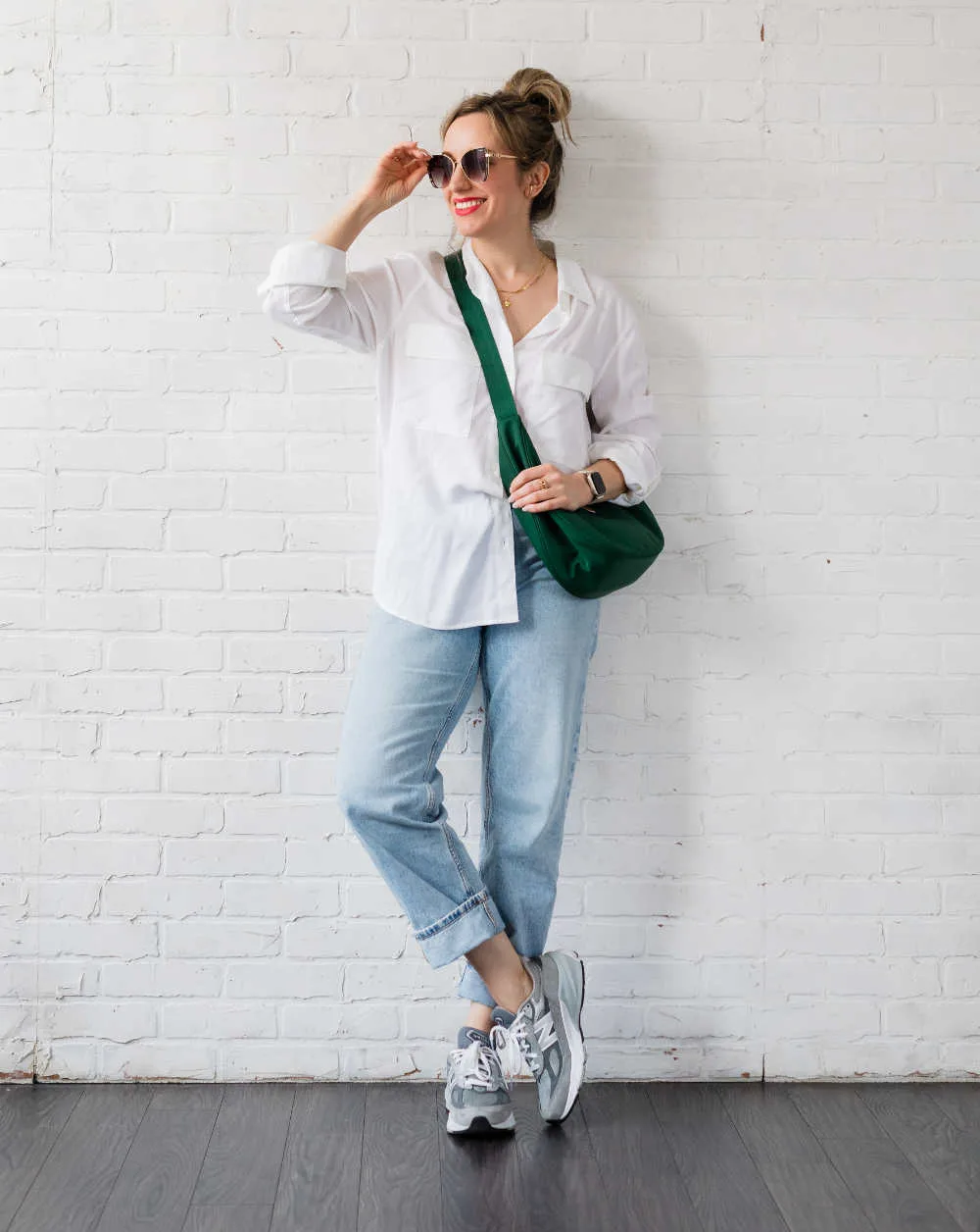 Woman wearing grey New Balance 990 V6 dad sneakers with rolled up faded straight leg jeans, a white shirt, green crossbody moon shaped bag and sunglasses.